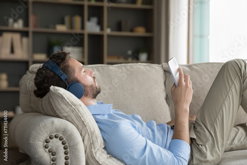 Peaceful young man relaxing alone on sofa with smartphone, listen quality sound through modern wireless headphones, spend carefree weekend leisure at home. On-line music streaming service apps user