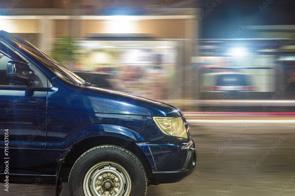 panning photography of a dark blue minibus driving fast at night in an urban area with shops
