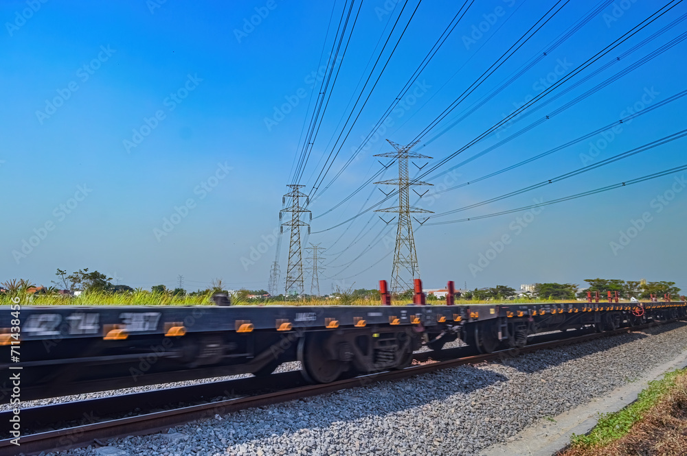 a series of flat-car trains speeding by against the backdrop of high-voltage power lines during a clear blue sky