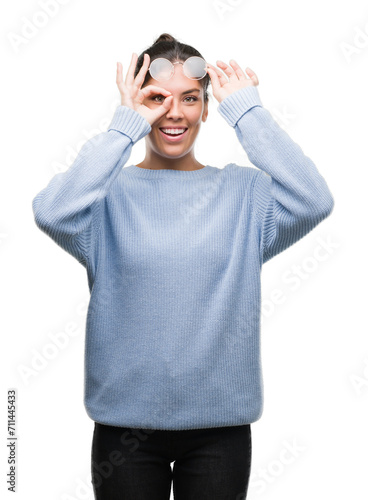 Young beautiful hispanic woman wearing a bun with happy face smiling doing ok sign with hand on eye looking through fingers