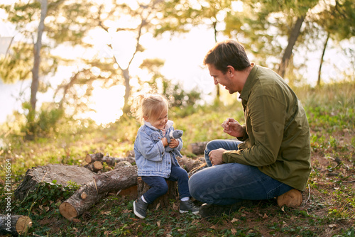 Laughing dad with a little daughter are sitting on stumps in a sunny meadow in the forest