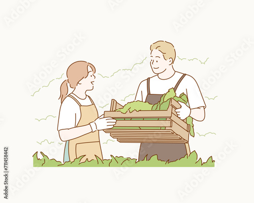 Smiling attractive young  farmer couple in apron standing, holding a wooden crate full of a variety of organic vegetables freshly harvested from an organic greenhouse farm. 