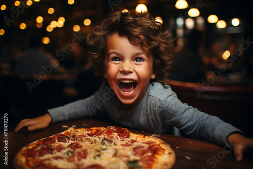  A happy laughing curly-haired boy at the table is eating fresh pizza, holding food in his hands. A joyful atmosphere in the pizzeria
