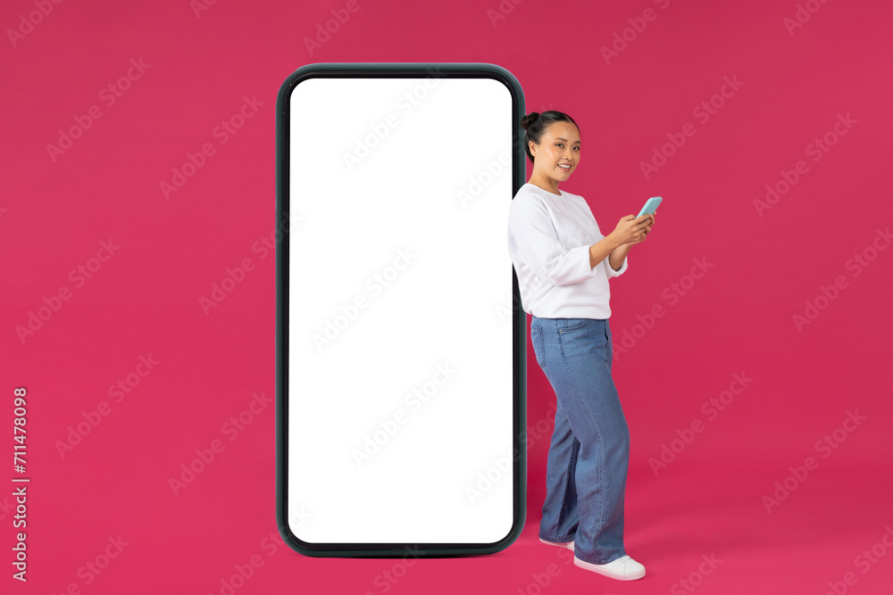 Great App. Asian Female Using Smartphone And Leaning At Big Blank Cellphone Great App. Asian Female Using Smartphone And Leaning At Big Blank Cellphone