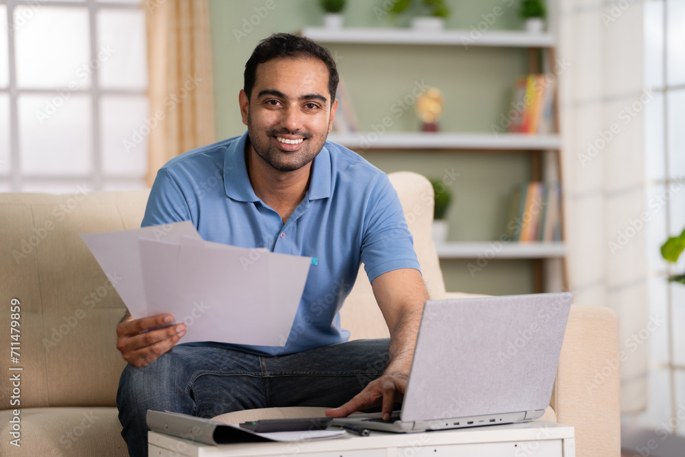 Happy Indian man checking financial documents or insurance bills looking at camera while working on laptop at home - concept of Budget tracking, Expense management and Contract review - obrazy, fototapety, plakaty 