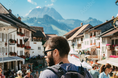 Young caucasian man tourist with beard and wearing sunglasses traveling with backpack exploring the european city Gruyères in Fribourg, Switzerland on summer season. SHOTLISTtravel. photo