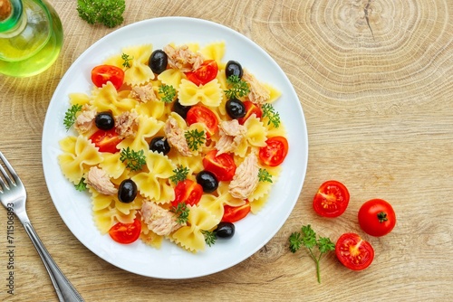 Farfalle pasta salad with canned tuna in olive oil, cherry tomatoes,black olives,parleys,olive oil and peppers on plate with wooden background.Healthy Italian summer salad.Top view.Copy space

