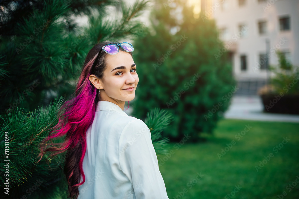 A beautiful high school student with purple hair smiles and looks at the camera. Portrait of a girl with glasses in the park on the background of the university A beautiful high school student with purple hair smiles and looks at the camera. Portrait of a girl with glasses in the park on the background of the university