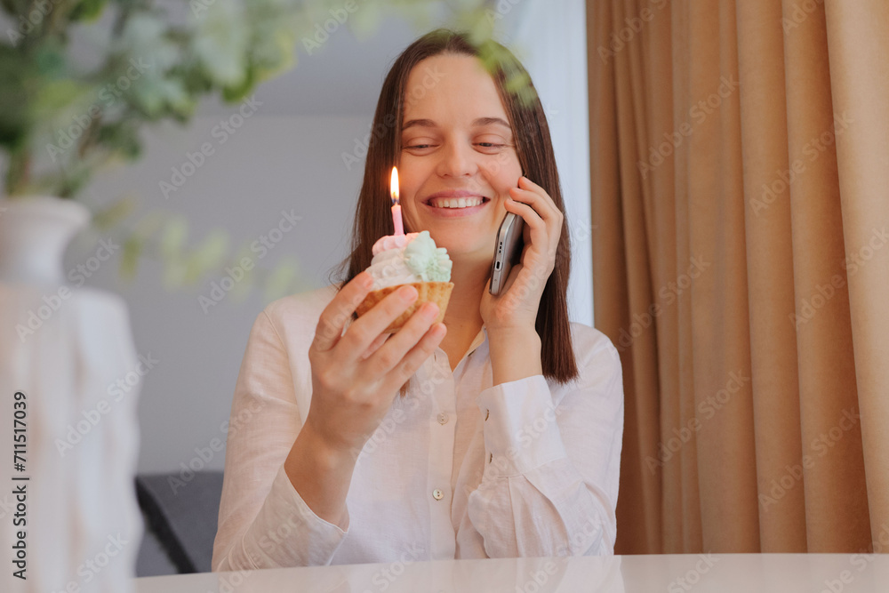 Smiling cheerful brown haired female in white shirt holding delicious sweet with candle talking phone with excited look smiling and communicating with her friend