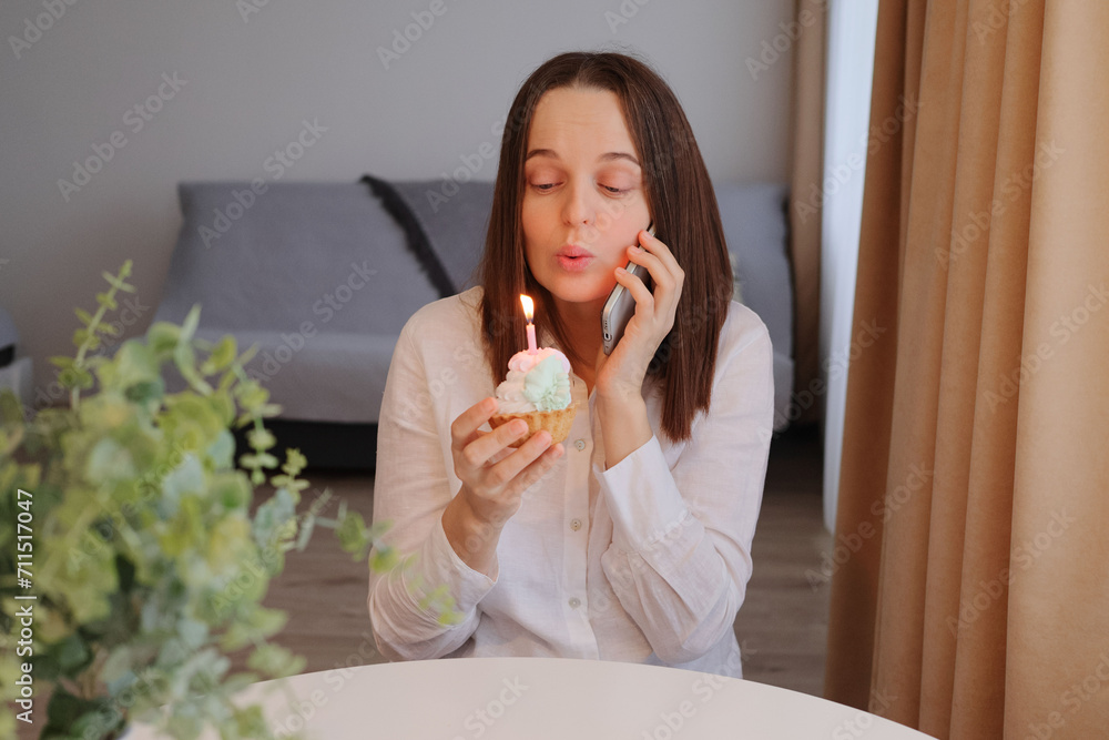 Funny dark haired woman wing white shirt sitting at home at table blowing candle on birthday cake and talking phone celebrating her birthday via call felling happy on holiday even alone Funny dark haired woman wing white shirt sitting at home at table blowing candle on birthday cake and talking phone celebrating her birthday via call felling happy on holiday even alone