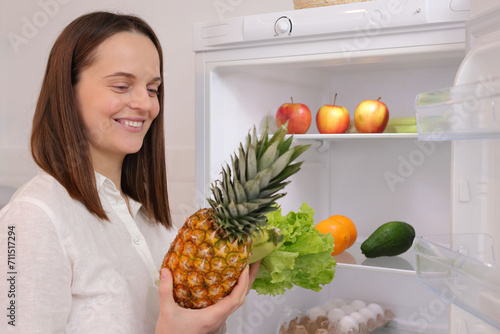 Smiling satisfied Caucasian woman standing near open refrigerator full of fruits and vegetables enjoying healthy eating talking pineapple for healthy snack from refrigerator photo