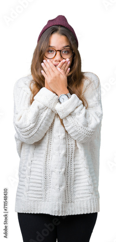 Young beautiful brunette hipster woman wearing glasses and winter hat over isolated background shocked covering mouth with hands for mistake. Secret concept.