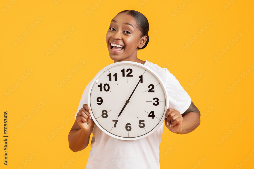 Cheerful Young African American Woman Showing Round Clock, Yellow Backdrop Cheerful Young African American Woman Showing Round Clock, Yellow Backdrop