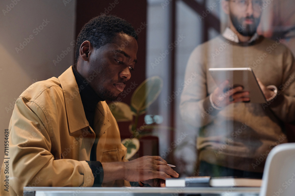 Black man in casual outfit working at desk in office, his caucasian colleague standing on background Black man in casual outfit working at desk in office, his caucasian colleague standing on background