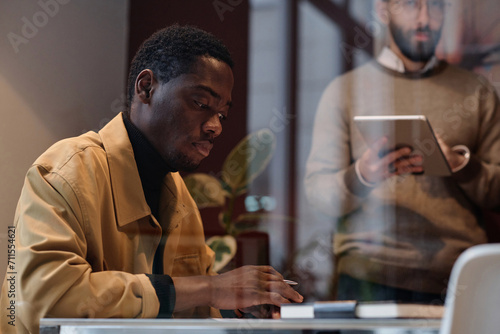 Black man in casual outfit working at desk in office, his caucasian colleague standing on background