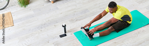 young african american man in stylish sportswear stretching on fitness mat in living room, banner