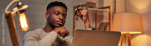 pensive african american freelancer holding eyeglasses looking at laptop in home office, banner