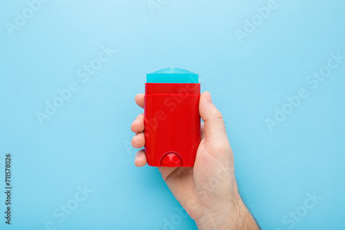 Young adult man hand holding and showing red plastic stick of armpit deodorant on light blue table background. Pastel color. Male daily beauty product. Closeup. Top down view. photo