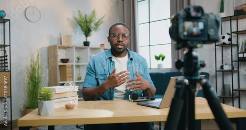 Attractive african man blogger in glasses filming new video for his audience with professional videocamera on home interior background photo