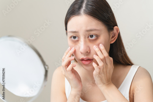 Bored, insomnia asian young woman, girl looking at mirror hand touching under eyes with problem of black circles or panda puffy, swollen and wrinkle on face. Sleepless, sleepy healthcare person. photo