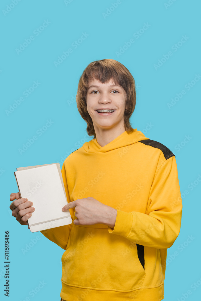 Young smiling teen boy holding notebook isolated on blue background, Teen boy's bright smile radiates as he holds a notebook with confidence. Young smiling teen boy holding notebook isolated on blue background, Teen boy's bright smile radiates as he holds a notebook with confidence.