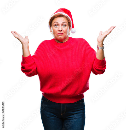 Atrractive senior caucasian redhead woman wearing christmas hat over isolated background clueless and confused expression with arms and hands raised. Doubt concept.