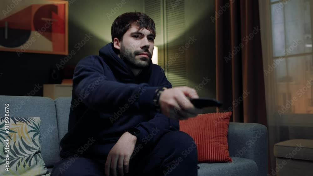 Man sitting at sofa at stylish apartment. Male relaxing after work day. Handsome male watching TV program. Man spending leisure watching series. Male changing TV channels with remote control. Man sitting at sofa at stylish apartment. Male relaxing after work day. Handsome male watching TV program. Man spending leisure watching series. Male changing TV channels with remote control.
