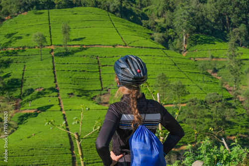 Blonde hair female cyclist in helmet looking at tea plantations and forest, rear view