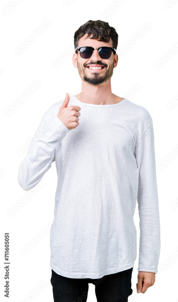 Young handsome man wearing sunglasses over isolated background doing happy thumbs up gesture with hand. Approving expression looking at the camera with showing success. Young handsome man wearing sunglasses over isolated background doing happy thumbs up gesture with hand. Approving expression looking at the camera with showing success.