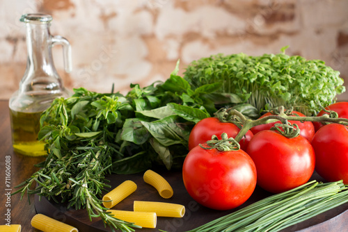 fresh ingredients, tomatoes, rosemary, oregano, parsley, chives, raw pasta dough, a board and wooden table, in the background olive oil in a container, blurred brick background