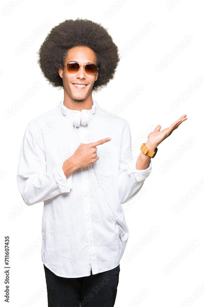 Young african american man with afro hair wearing sunglasses and headphones amazed and smiling to the camera while presenting with hand and pointing with finger. Young african american man with afro hair wearing sunglasses and headphones amazed and smiling to the camera while presenting with hand and pointing with finger.