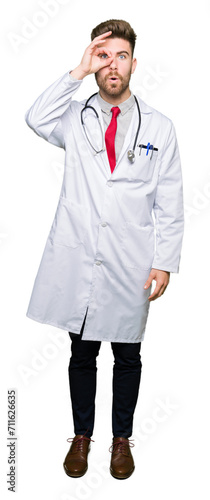 Young handsome doctor man wearing medical coat doing ok gesture shocked with surprised face, eye looking through fingers. Unbelieving expression.