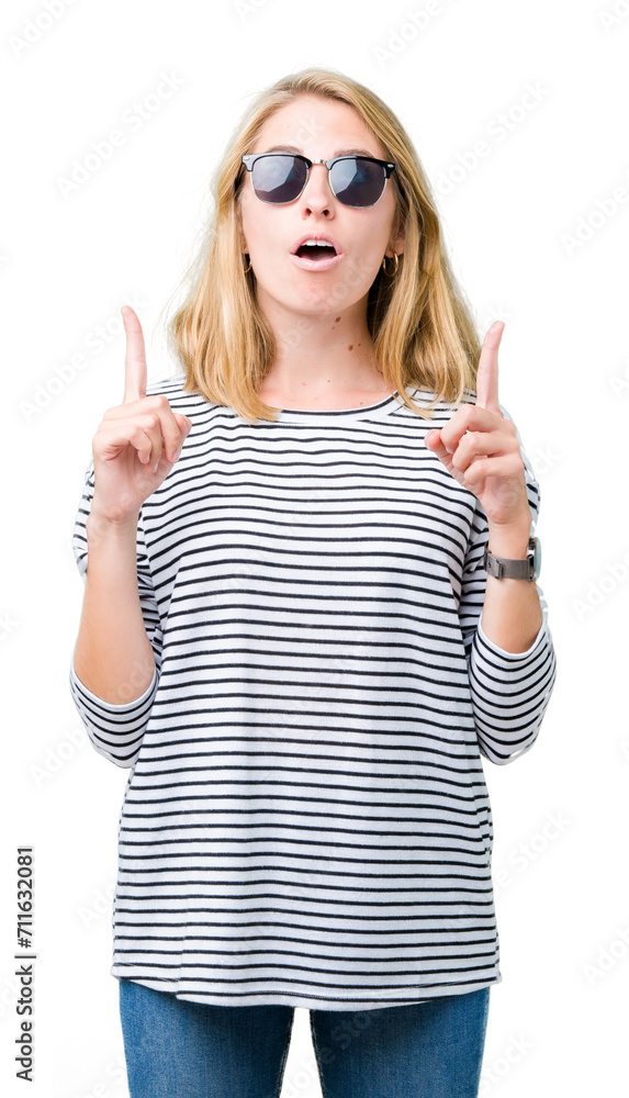 Beautiful young woman wearing sunglasses over isolated background amazed and surprised looking up and pointing with fingers and raised arms. Beautiful young woman wearing sunglasses over isolated background amazed and surprised looking up and pointing with fingers and raised arms.