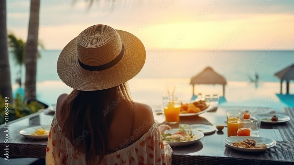 Dinner with sea view in luxury hotel. Woman in straw hat near swimming pool, eating food and enjoy ocean view. Dinner table on tropical vacation. Back view. Concept of travel, holidays, weekend.