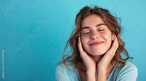 Cheerful female smiling for camera and clasping hands under cheek while pretending to sleep against blue background photo
