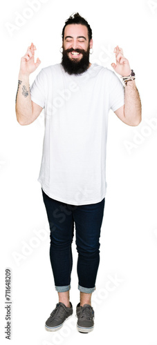 Young hipster man with long hair and beard wearing casual white t-shirt smiling crossing fingers with hope and eyes closed. Luck and superstitious concept.