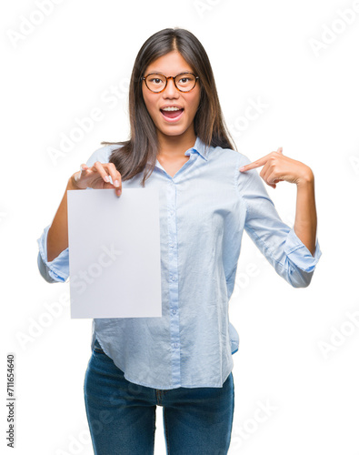 Young asian woman over isolated background holding blank paper with surprise face pointing finger to himself