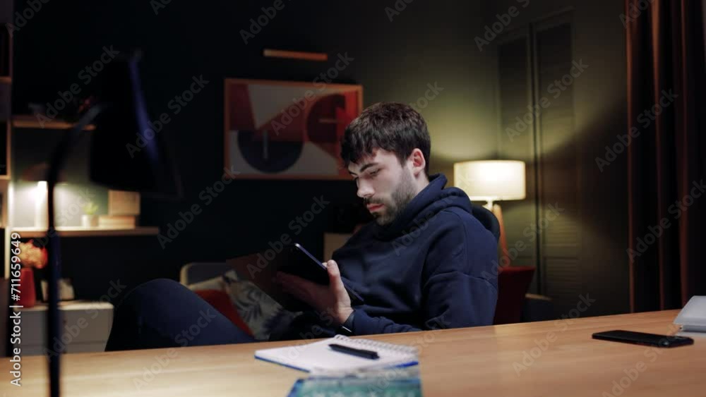 Man sitting in chair and looking for information in Internet. Male looking at tablet screen. Man sitting at home at table. Male working at home distantly. Man putting tablet at table. Man sitting in chair and looking for information in Internet. Male looking at tablet screen. Man sitting at home at table. Male working at home distantly. Man putting tablet at table.