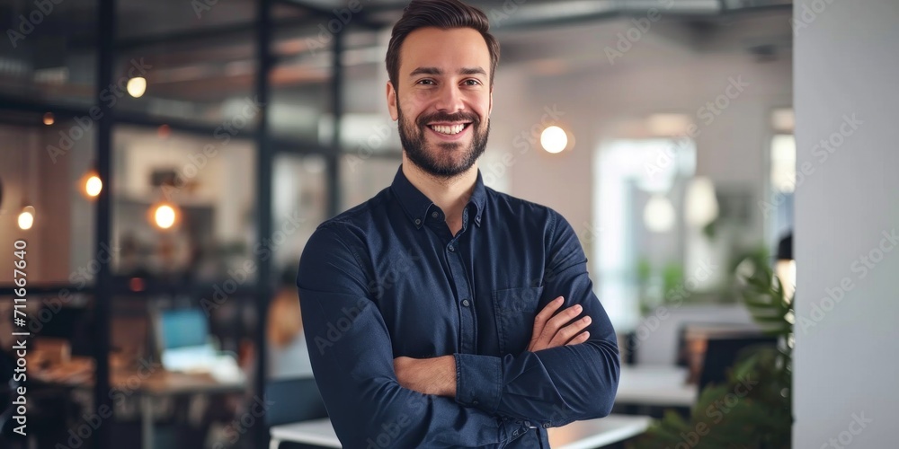 Handsome smiling young fashion designer stands, arms crossed, modern office in the background Handsome smiling young fashion designer stands, arms crossed, modern office in the background