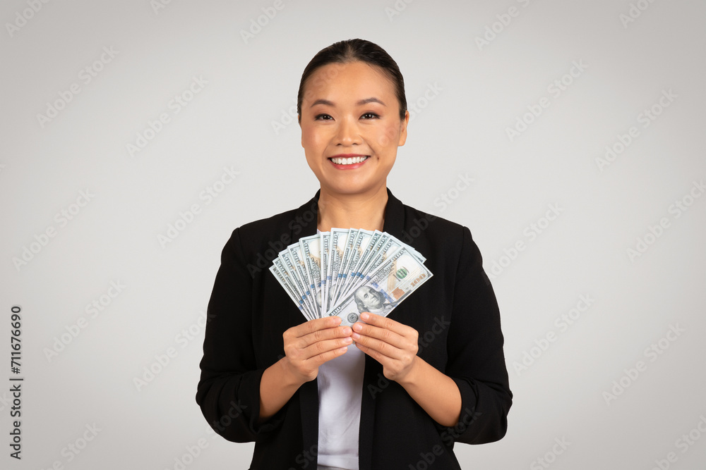 A radiant Asian businesswoman in professional attire beams as she offers a fan of hundred-dollar bills A radiant Asian businesswoman in professional attire beams as she offers a fan of hundred-dollar bills
