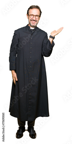 Middle age priest man wearing catholic robe smiling cheerful presenting and pointing with palm of hand looking at the camera.