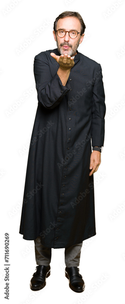 Middle age priest man wearing catholic robe looking at the camera blowing a kiss with hand on air being lovely and sexy. Love expression.