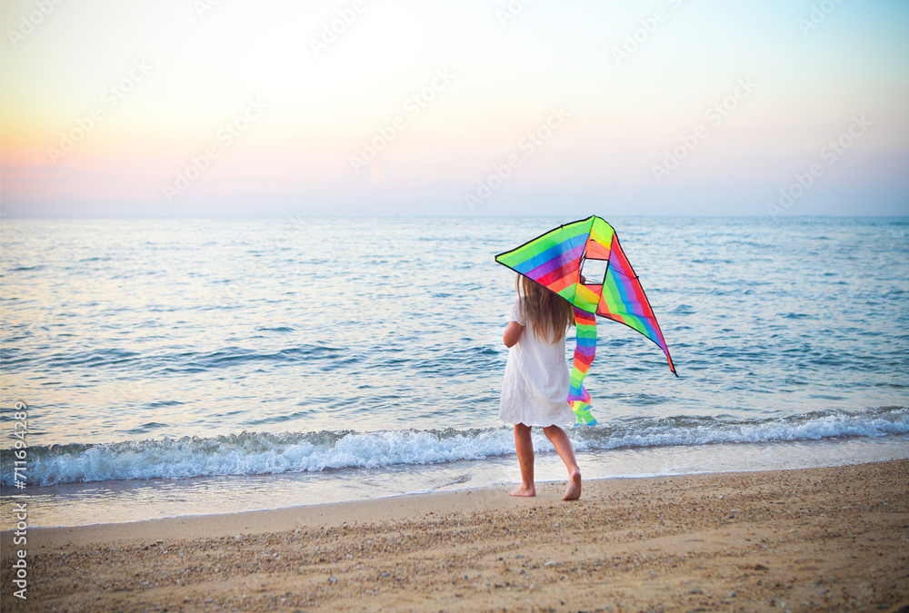 Little running girl with flying kite on beach at sunset