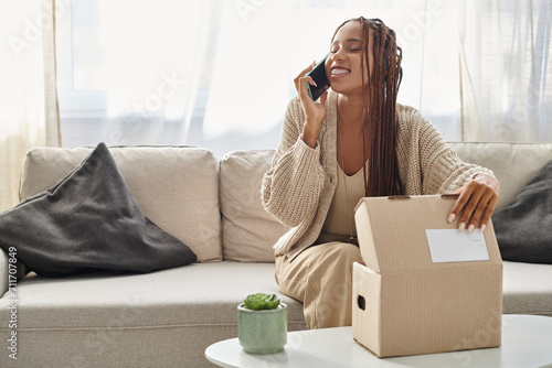 joyful attractive african american woman in comfy homewear talking by phone with hand on parcel photo