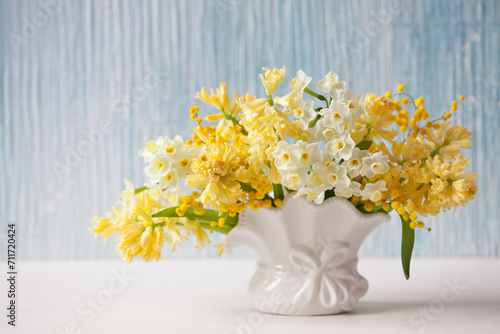 Spring bouquet of yellow hyacinths, daffodils, mimosa in a vase on a table, blue background wall. Closeup, blur, soft focus.