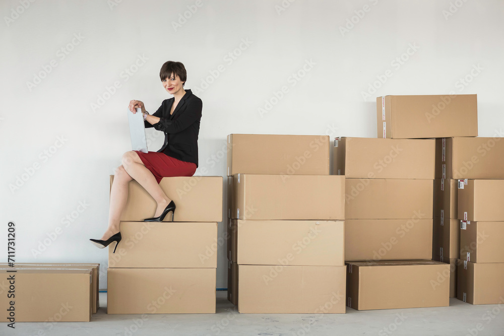 Female owner of a startup sits on top of moving boxes in the office with business plan in hand. Female owner of a startup sits on top of moving boxes in the office with business plan in hand.