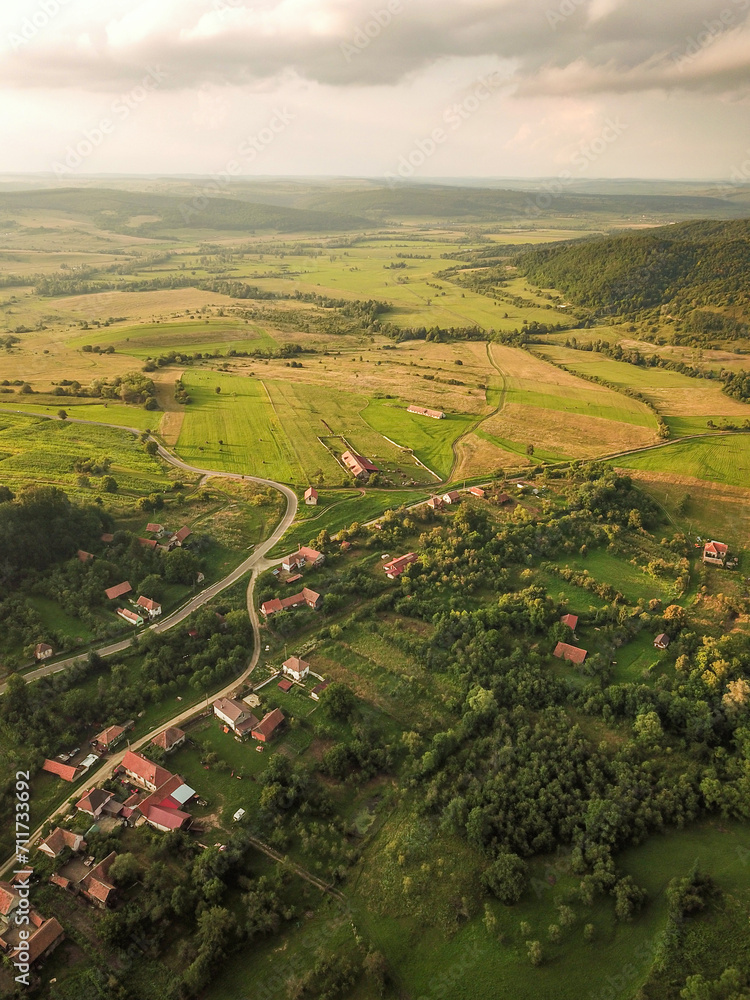 Aerial drone flight above a rural region in Transylvania at sunset. Grassy hills, orchards, agricultural fields, meadows, storm clouds and sunlight create a beautiful countryside landscape. Romania