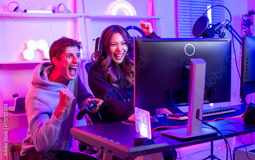 Couple or friends have shocking, exciting face with fun, using joystick to compete and play games together, sitting in room at home with neon decorated light. Game online streamer Concept.