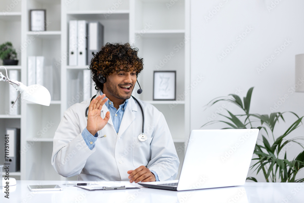 Engaging male Hindu doctor in white coat with headset waving at the camera, providing a warm virtual consultation from his well-lit office. Engaging male Hindu doctor in white coat with headset waving at the camera, providing a warm virtual consultation from his well-lit office.