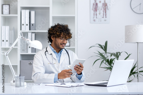 A professional young doctor in a white coat attentively uses a digital tablet in a bright medical office.